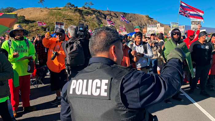 A police officer gestures at demonstrators blocking a road at the base of Hawaii's tallest mountain, Monday, July 15, 2019, in Mauna Kea, Hawaii.
