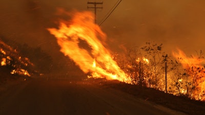 In this Nov. 9, 2018 file photo Wind-driven flames from a wildfire race up a slope and cross the road in Malibu, Calif. known as the Woolsey Fire, it has consumed tens of thousands of acres and destroyed multiple homes. Southern California Edison announced in its quarterly earnings report that its equipment probably caused the November 2018 Woolsey Fire that raged from north of Los Angeles through Malibu to the sea, killing three people and burning more than 1,600 homes and other buildings.
