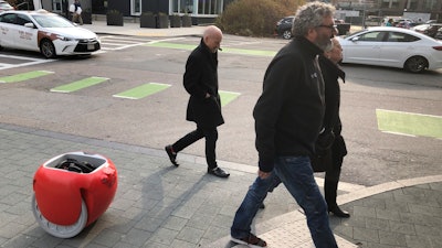 Piaggio Fast Forward CEO Greg Lynn, center, is followed by his company's Gita carrier robot as he crosses a street on Monday, Nov. 11, in Boston. The two-wheeled machine is carrying a backpack and uses cameras and sensors to track its owner.