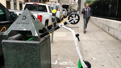 In this photo taken May 14, 2018, a man walks past an electric scooter that dumped into a trash can in San Francisco.