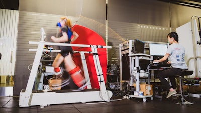 Graduate student Delaney Miller runs on a treadmill aided by the ankle exoskeleton emulator. Fellow graduate student Guan Rong Tan controls the emulator and monitors Miller's gait and respiration.