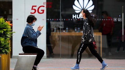 A woman wearing a face mask to help curb the spread of the coronavirus browses her smartphone as a masked woman walks by the Huawei retail shop promoting it 5G network in Beijing on Oct. 11, 2020. Chinese leaders are meeting to formulate an economic blueprint for the next five years that is expected to emphasize development of semiconductors and other technology amid a feud with Washington that is cutting off access to U.S. components for China's fledgling tech industries.
