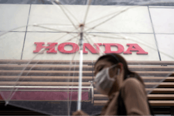 A woman walks in the rain near the logo of Honda Motor Company Friday, May 13, 2022, in Tokyo. Honda’s fiscal fourth quarter profit slipped to almost half of what the Japanese automaker earned the previous year amid headwinds of supply shortages and rising raw material costs.