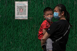 A woman wearing a face mask carries a masked child stands against a board displaying a health QR code on Wednesday, May 18, 2022, in Beijing. Angry bank customers who traveled to a city in central China attempting to retrieve their savings from troubled rural banks were stopped in their tracks by a common technology: a QR code.