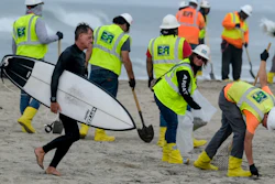 A surfer walks by as workers in protective suits continue to clean the contaminated beach in Huntington Beach, Calif., on Oct. 11, 2021.