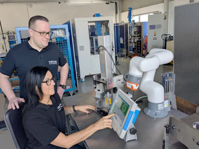 Two employees operating a white collaborative robot in an industrial manufacturing facility with blue machinery in the background