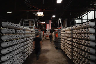 Industrial warehouse interior with stacked cylindrical material rolls lining both sides of a central aisle, American flag visible overhead