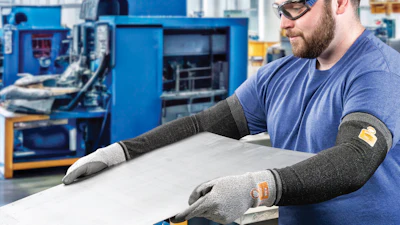 Factory worker in safety glasses and protective gloves inspecting white material at workstation with blue industrial machinery in background