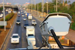 Traffic enforcement camera mounted on pole overlooking busy multi-lane highway with vehicles and urban infrastructure