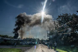 Dark smoke plume rises from industrial fire in residential area under bright sunlight with parked vehicles and onlookers nearby