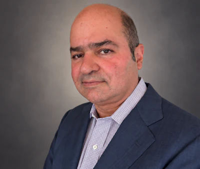Professional headshot of a middle-aged man wearing a dark blazer and light purple checkered shirt against a neutral gray background