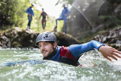 Man canyoning in turquoise river wearing helmet and blue harness with group in background