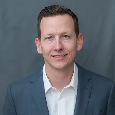 Professional headshot of a smiling man in a navy blazer and white shirt against a gray background
