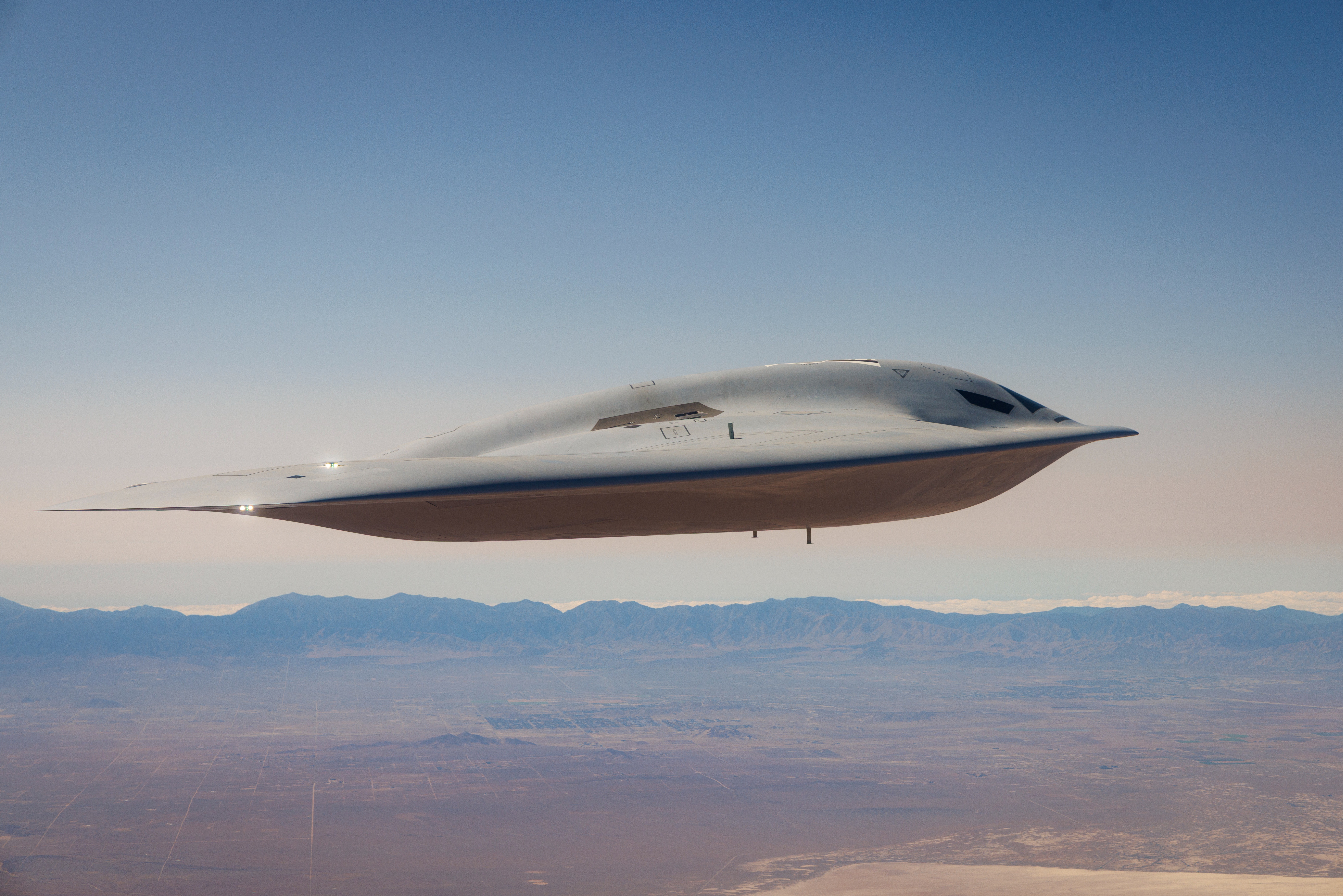 Futuristic silver aircraft with streamlined design flying over desert landscape and distant mountains