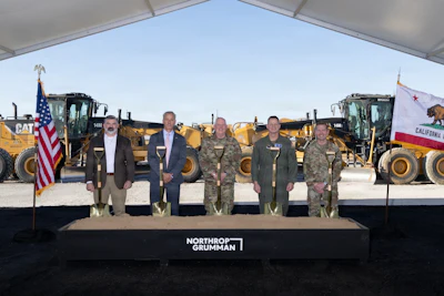 Six officials stand at podium with Northrop Grumman logo, flanked by American and California flags, with heavy equipment in background