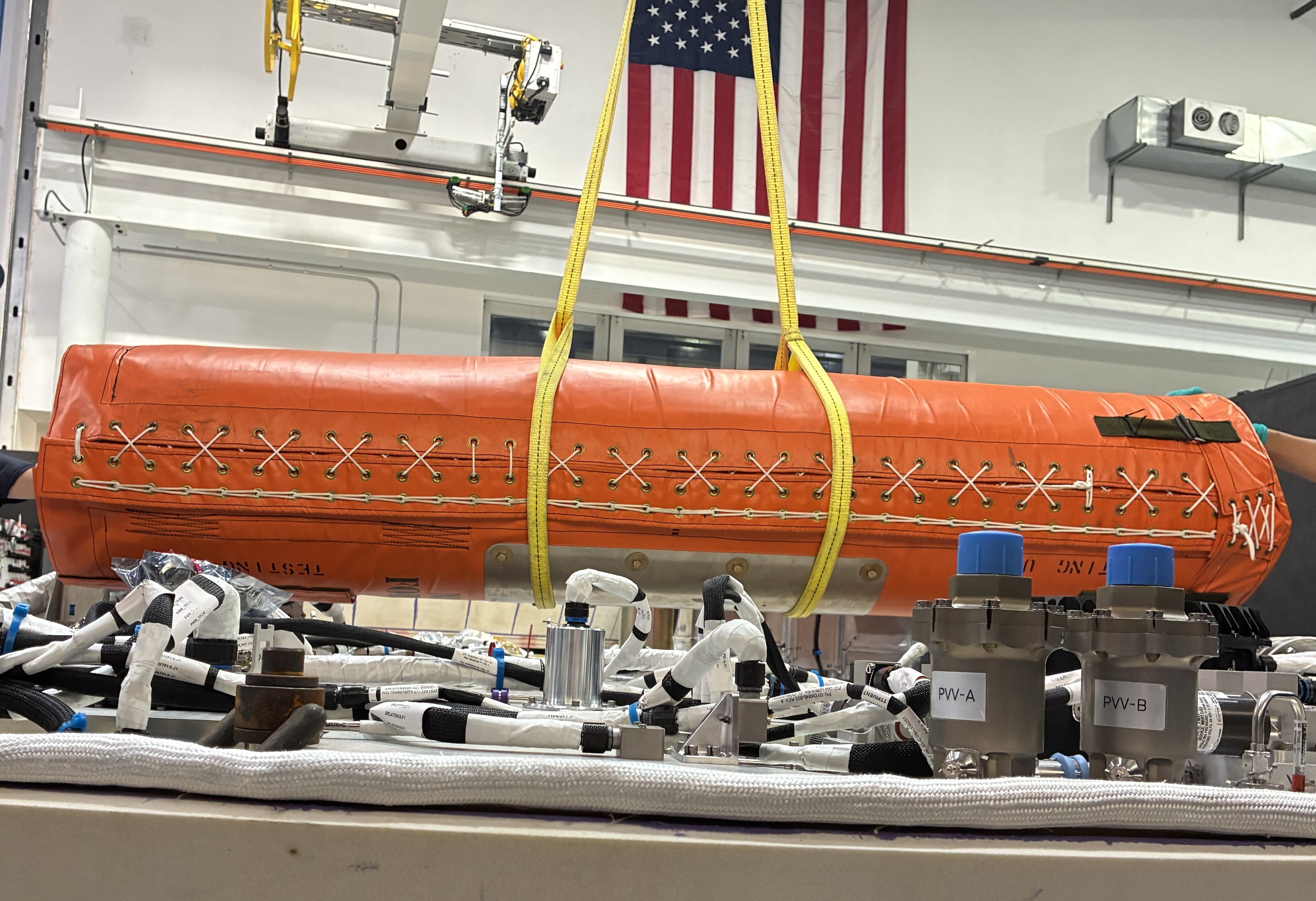 Orange cylindrical aerospace equipment suspended by yellow straps in industrial workshop with American flags