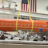 Orange cylindrical aerospace equipment suspended by yellow straps in industrial workshop with American flags