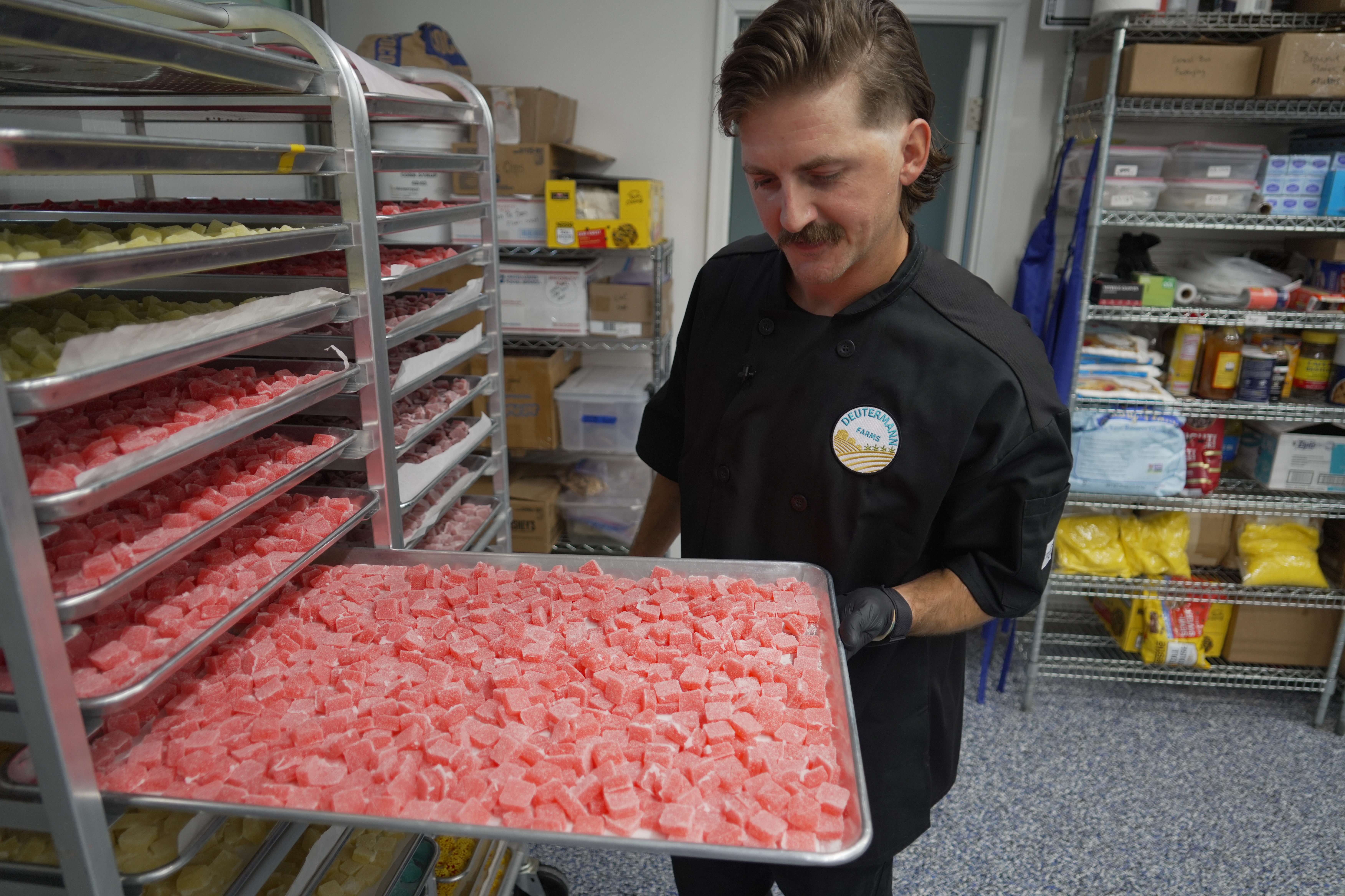 Worker holding tray of pink gummy candies in commercial kitchen with shelving and inventory