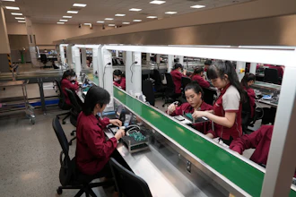 Electronics factory workers in maroon uniforms assembling components at industrial workstations in a bright manufacturing facility.
