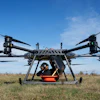 Large agricultural drone with orange payload container hovering above dry field under clear blue sky