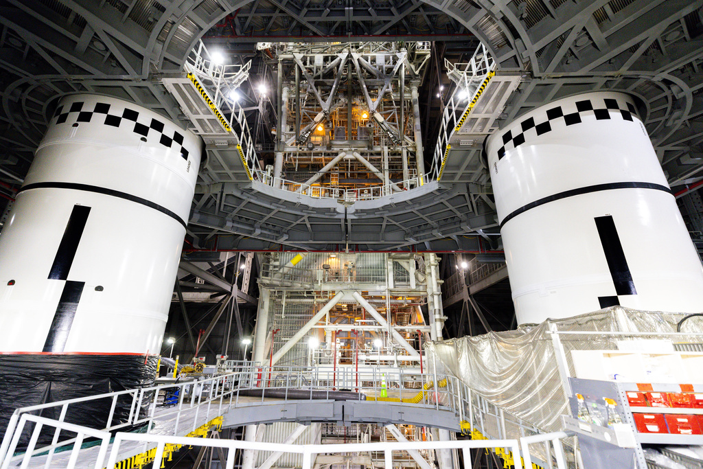 Two white spacecraft modules with black checkered docking targets in assembly facility with metal framework and equipment overhead.