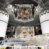 Two white spacecraft modules with black checkered docking targets in assembly facility with metal framework and equipment overhead.
