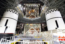 Two white spacecraft modules with black checkered docking targets in assembly facility with metal framework and equipment overhead.