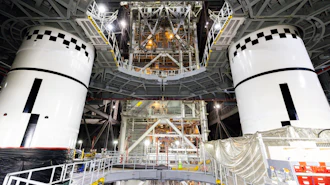 Two white spacecraft modules with black checkered docking targets in assembly facility with metal framework and equipment overhead.