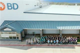 Large group of BD company employees standing together outside a modern industrial facility with white and teal metal roofing.