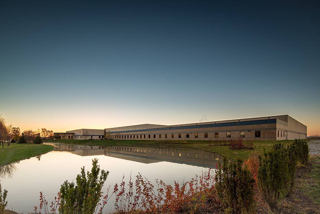 Modern industrial building complex reflected in calm water at dusk with autumn vegetation in foreground
