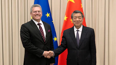 Two officials in formal business attire shake hands at a diplomatic meeting with EU and Chinese flags in the background.