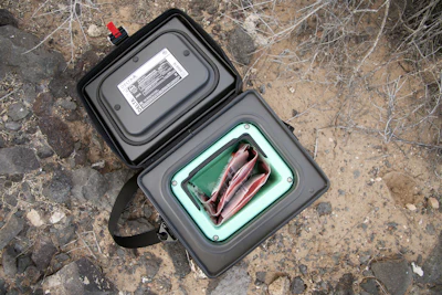 Open black portable tool organizer with mint green interior compartments containing red tools and accessories on dirt ground