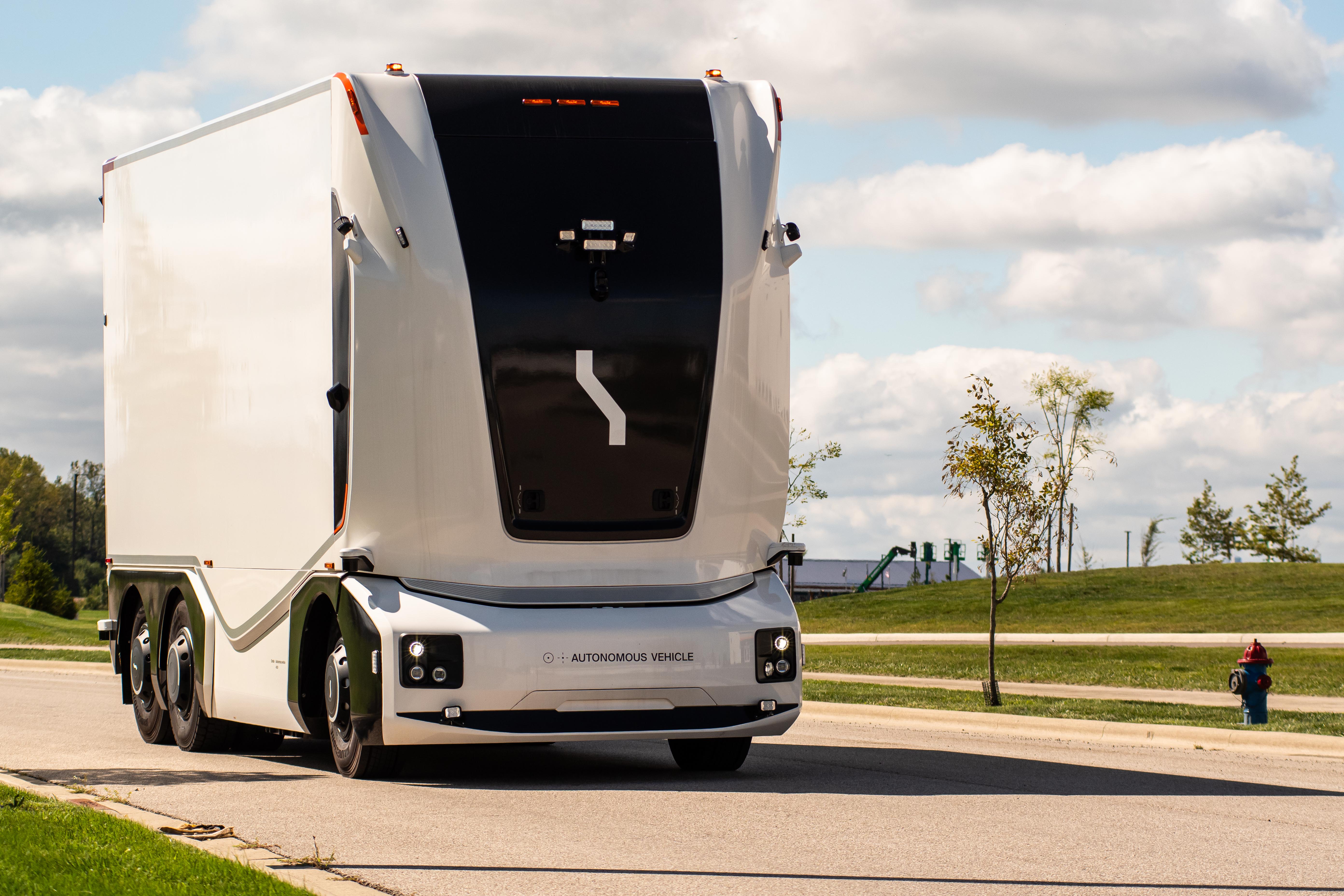 White autonomous semi-truck with black cabin parked on a paved road with green fields and cloudy sky in background