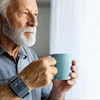 Senior man with gray beard holding a teal mug while looking out a window