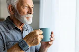 Senior man with gray beard holding a teal mug while looking out a window
