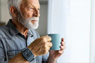 Senior man with gray beard holding a teal mug while looking out a window