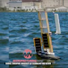 Autonomous solar-powered sailboat with white sails on blue water near waterfront buildings