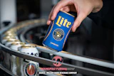 Hand holding blue Miller Lite beer can above industrial manufacturing equipment