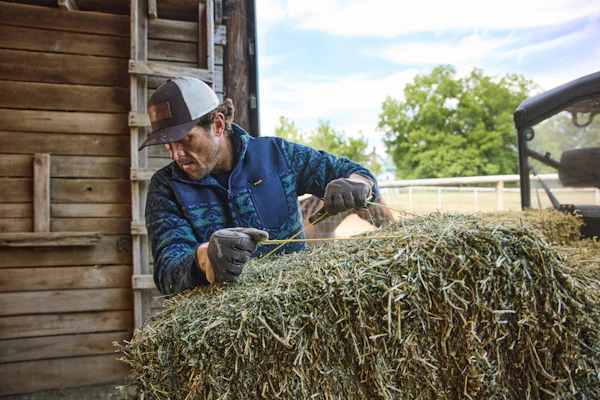 Farmer in blue jacket and cap handling hay bales near a wooden barn and truck