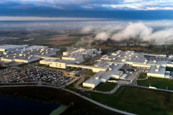 Aerial view of large industrial manufacturing complex with warehouse facilities, parking areas, and surrounding countryside landscape under cloudy sky