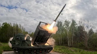Military MLRS launcher firing a long-range missile in a forest clearing with smoke and flames visible