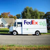 FedEx delivery truck parked on residential street in front of white house with autumn trees