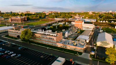 Aerial view of brick industrial complex with parking lot, green spaces, and surrounding cityscape
