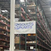 Industrial warehouse interior with tall storage racks, branded signage including Goulds Pumps and Rexnord logos, pallets, and orange safety markers on concrete floor.