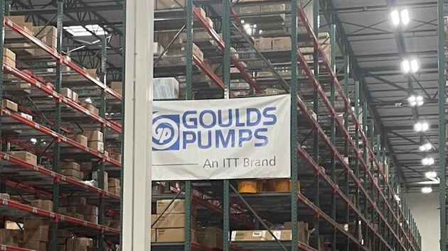 Industrial warehouse interior with tall storage racks, branded signage including Goulds Pumps and Rexnord logos, pallets, and orange safety markers on concrete floor.