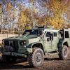 Tactical armored military vehicle with open door parked outdoors against autumn trees
