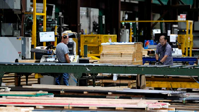 Workers processing lumber at an industrial woodworking facility with yellow machinery and green structural beams.