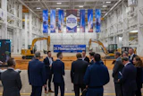 Corporate event in industrial warehouse with blue banner reading 'Future Home of 24 Hadrian Machines' and yellow excavators