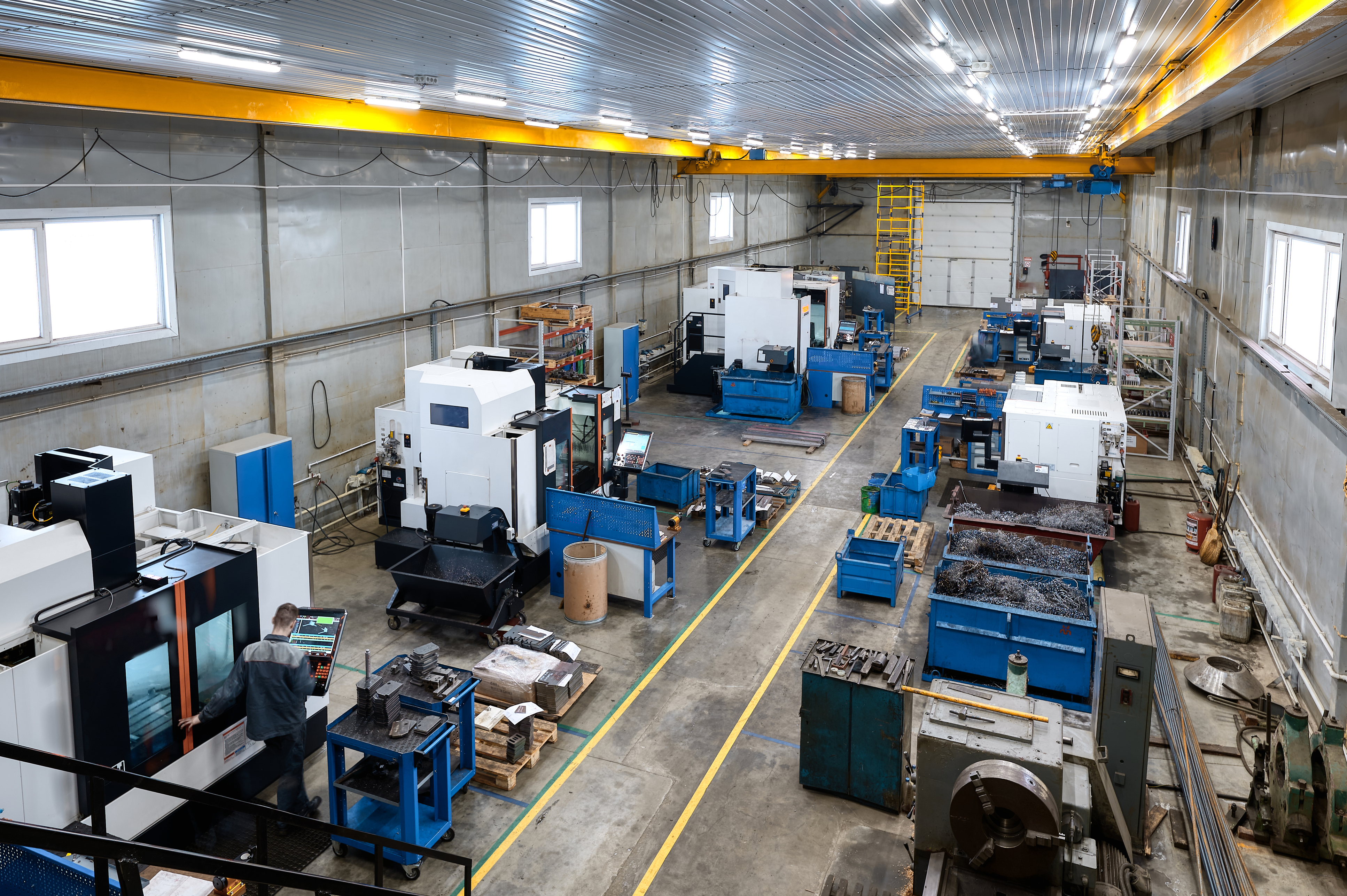 Industrial manufacturing facility with CNC machines, blue workbenches, and overhead yellow crane system in warehouse space