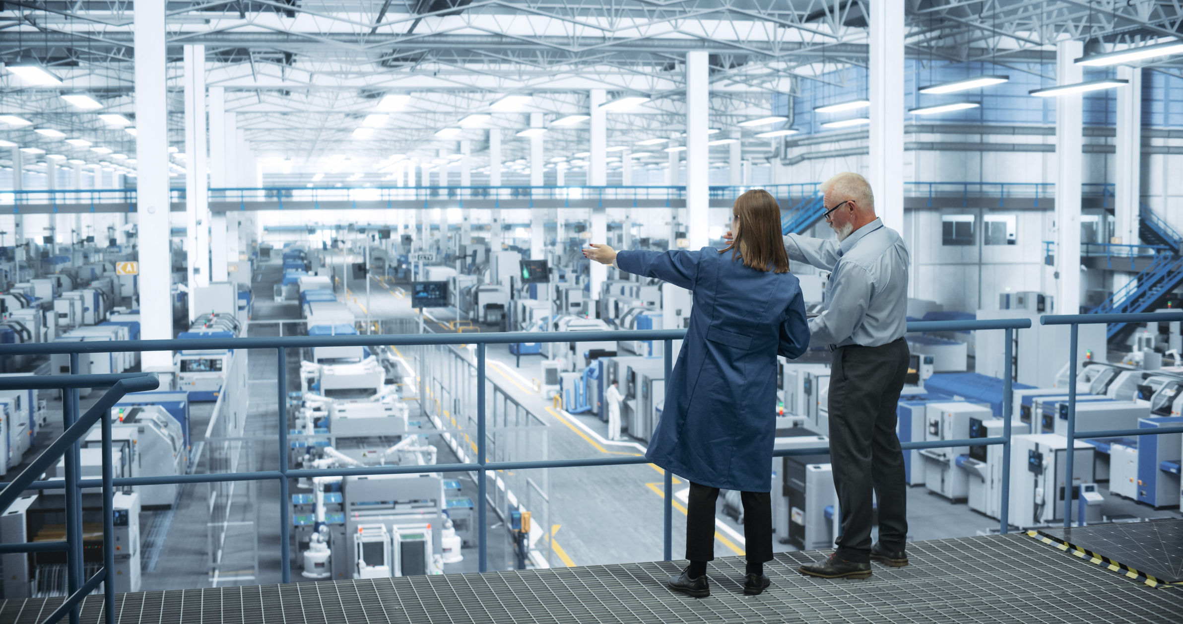 Two professionals observe a large industrial manufacturing facility from an elevated platform with expansive windows.